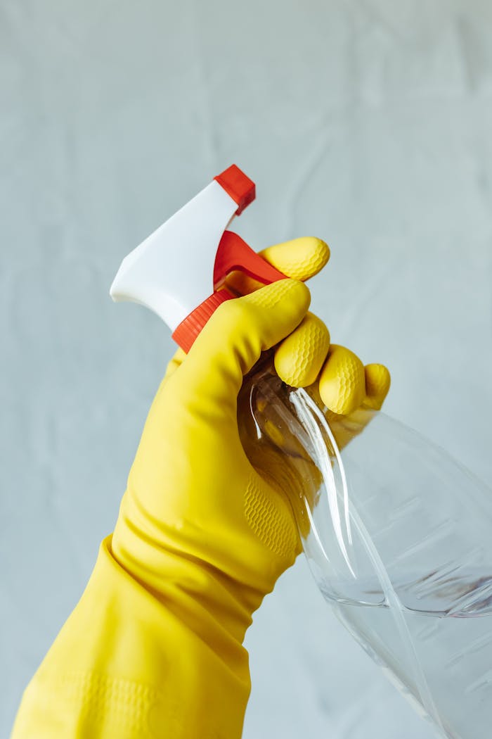 services-01 A hand wearing a yellow rubber glove holds a red and white spray bottle against a neutral background.